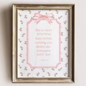 Pode incluir: Uma impressão emoldurada com uma moldura dourada. A impressão apresenta um fundo com um padrão floral rosa e branco. O texto "She is more precious than rubies, nothing you desire can compare with her" é exibido em uma moldura rosa com um laço.