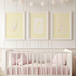 Può includere: Tre stampe artistiche incorniciate per la cameretta. Due stampe presentano un'illustrazione di un'oca bianca su sfondo giallo con un bordo a margherite. La stampa centrale mostra la lettera "Z" su sfondo giallo. Una culla bianca con biancheria da letto rosa è in primo piano.