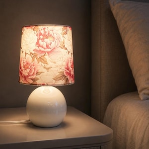 May include: A table lamp with a spherical white ceramic base and a tapered cylindrical lampshade featuring a floral pattern of pink and white peonies with green leaves. The lamp emits a warm glow and includes a white power cord.