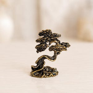 May include: A small, antique brass-colored metal figurine shaped like a windswept bonsai tree. The sculpture features a textured, gnarled trunk and layered, rounded foliage clusters, all resting on a solid, uneven base with a weathered finish.