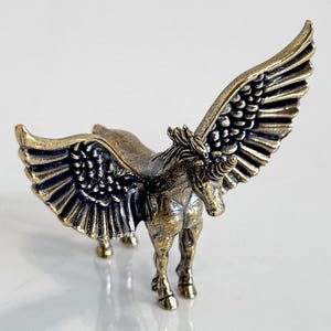 May include: A bronze-colored figurine of a winged horse, or Pegasus, with outstretched wings. The sculpture has detailed feathering on the wings and a flowing mane. The horse stands on four legs, with a detailed body.
