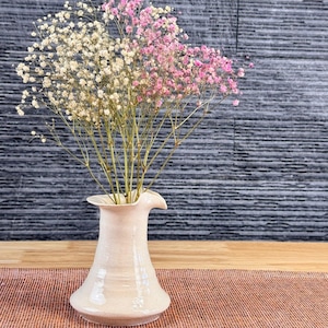 May include: A white ceramic vase with a unique spout design, holding a bouquet of white and pink baby's breath flowers. The vase is sitting on a brown woven placemat.