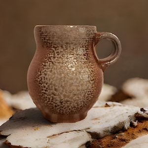 以下が含まれることがあります： 岩の形成の上に置かれた、質感のある表面を持つ茶色のセラミックマグカップ。マグカップは丸みを帯びたボディとハンドルを持っています。