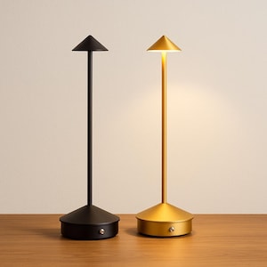 May include: Two modern table lamps. One is black, the other is gold. Each lamp has a conical shade and a cylindrical stem. The bases are round. The gold lamp is illuminated, casting a warm glow.