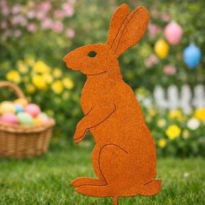 Puede incluir: Una estaca de jardín de metal naranja oxidado con forma de conejito de Pascua. El conejito está en posición sentada con orejas largas y un ojo recortado. Una cesta de huevos de Pascua coloridos y una valla blanca están en el fondo.