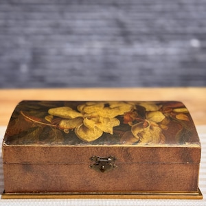 May include: A wooden box with a hinged lid and a floral design. The box has a brown background with a floral pattern in shades of yellow, orange, and green. The box has a brass latch and a gold trim around the edges.