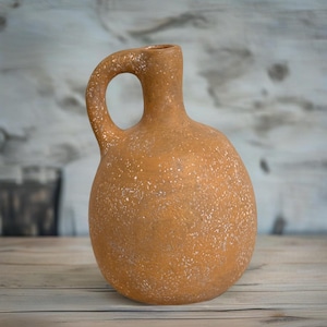 May include: A rustic brown ceramic pitcher with a speckled finish and a rounded body. The pitcher has a single handle and a narrow opening.