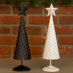 Puede incluir: Dos árboles de Navidad decorativos de metal. Uno es negro con una estrella en la parte superior, el otro es blanco con una estrella en la parte superior. Ambos tienen un patrón de diamantes texturizado y se asientan sobre una base redonda. El fondo es una pared de ladrillos.