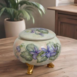 May include: A ceramic trinket box with a lid, decorated with a floral design of purple violets and green leaves. The box has three gold-colored feet.