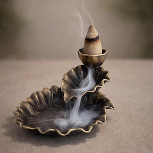May include: A bronze-colored incense burner with a leaf design. A lit incense cone sits in the top bowl, emitting a stream of white smoke that cascades down into the lower leaf-shaped dish. The background is a neutral, blurred tone.