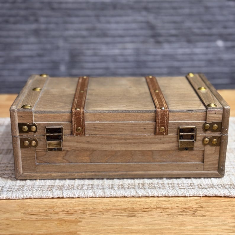 May include: A wooden box with a distressed finish and brown leather straps. The box has brass hardware and is closed.