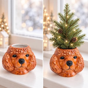 Pode incluir: Um vaso de cerâmica em forma de cabeça de poodle castanho com pelo texturizado, olhos pretos e nariz preto. O vaso é mostrado vazio e também contendo um pequeno pinheiro verde decorado com pequenas luzes brancas e pinhas castanhas.