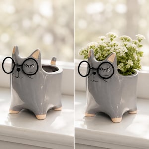 Op de afbeelding: Grijze keramische katvormige plantenbak met zwarte bril en gesloten ogen. De pot heeft een kleine staart en beige poten. De plantenbak is leeg of gevuld met witte bloemen.