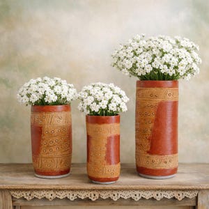May include: Three cylindrical vases with a reddish-brown and tan color scheme, each filled with white flowers. The vases have a textured, patterned design. They are arranged on a wooden table with a decorative edge.