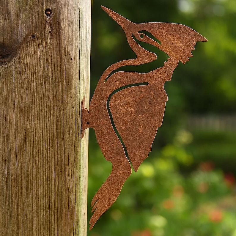 K&ouml;nnte beinhalten: Rostbraune Metallskulptur eines Spechts, befestigt an einem Holzpfosten. Der Vogel ist im Profil dargestellt, mit einem langen Schnabel und detaillierten Federn. F&uuml;r die Gartendekoration im Au&szlig;enbereich.