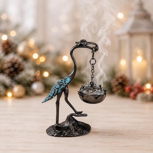 Puede incluir: Una figura de grulla de metal negro sobre una base, con detalles en turquesa en las alas. La grulla sostiene una cadena colgante con un quemador de incienso en forma de loto, del cual emana un flujo vertical de humo blanco.