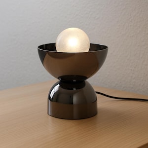 May include: A modern, black table lamp with a spherical, frosted glass shade. The lamp has a two-part, hourglass-shaped base and sits on a wooden surface. The lamp is plugged into an electrical outlet.