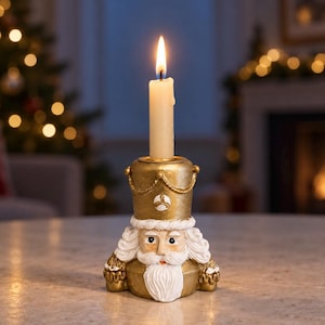 Puede incluir: Un portavelas con forma de cascanueces en color dorado y blanco que presenta un rostro esculpido con barba y bigote blancos, luciendo un sombrero dorado con un detalle de cuerda. Una vela cónica de color crema está insertada arriba, encendida.