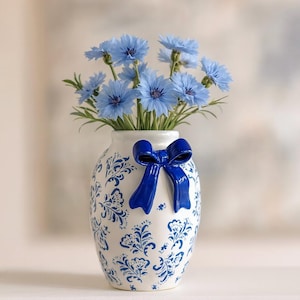 May include: A white ceramic vase with blue floral patterns and a 3D blue bow. The vase holds a bouquet of light blue flowers with green stems and leaves. The vase is a decorative item.
