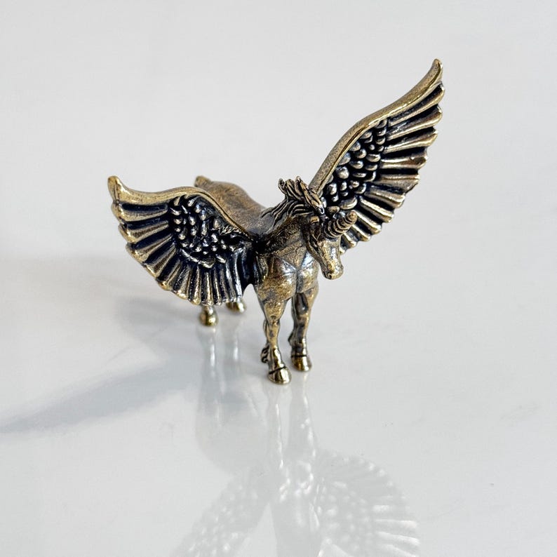 May include: A bronze-colored figurine of a winged horse, or Pegasus, with outstretched wings. The sculpture has detailed feathering on the wings and a flowing mane. The horse stands on four legs, with a detailed body.