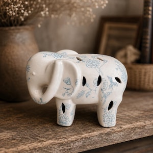 May include: A white ceramic elephant figurine with blue floral patterns. The elephant has a trunk, ears, and four legs. It has several circular cutouts on its body. The figurine is placed on a wooden surface.