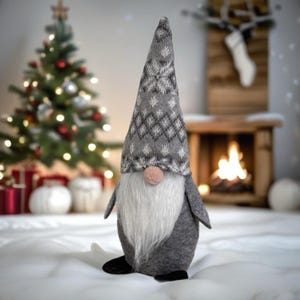 Peut inclure: Figurine de gnome de Noël avec une longue barbe blanche, un nez rose et un chapeau gris avec un motif de flocon de neige. Le gnome porte un corps gris et des chaussures noires. Un sapin de Noël et une cheminée sont en arrière-plan.