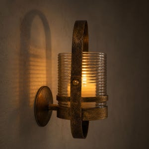 May include: A wall-mounted sconce featuring a cylindrical, ribbed clear glass shade held by a distressed bronze-toned metal frame with a vertical handle-like strap and a circular base plate, illuminated by a warm light source from within.