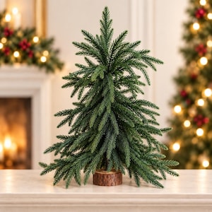 May include: A small artificial evergreen tree with dark green, needle-like foliage arranged in layered branches. The tree is supported by a circular, natural-toned wooden base with visible bark texture around the outer edge of the stand.