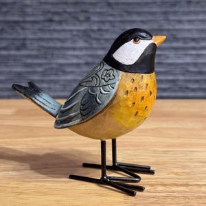 May include: A decorative bird figurine with a black head, white cheeks, and a yellow chest. The bird has a blue and gray wing with a floral design and stands on black metal legs. The beak is orange, and the bird is made of wood.