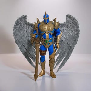 Puede incluir: Una figura de acción azul y dorada con grandes alas plateadas. La figura sostiene una espada en su mano derecha y tiene un casco en la cabeza.