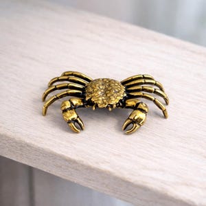 May include: A detailed, golden-colored crab figurine with textured detailing on its shell and claws. The crab has multiple legs and is positioned on a light-colored surface. This decorative item could be used as a cabinet pull or a decorative accent.