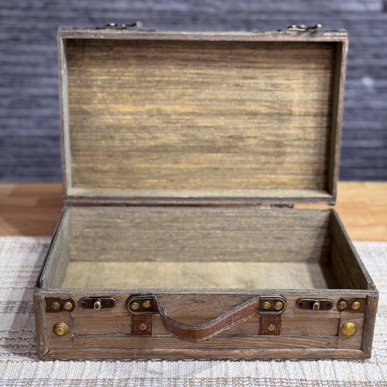 May include: A wooden suitcase with a brown leather handle and brass accents. The suitcase is open and empty.