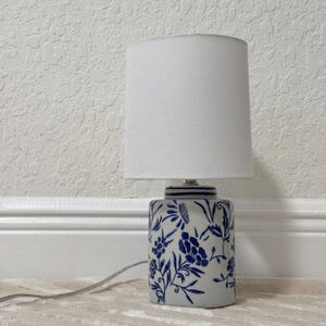 May include: A small table lamp with a white lampshade and a ceramic base. The base is white with a blue floral pattern. The lamp is plugged into an electrical outlet.