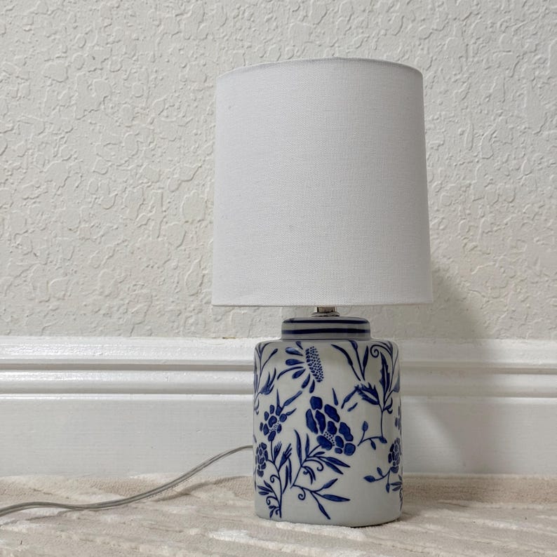 May include: A small table lamp with a white lampshade and a ceramic base. The base is white with a blue floral pattern. The lamp is plugged into an electrical outlet.