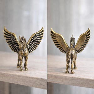 Puede incluir: Una figura de Pegaso de color bronce con las alas extendidas. La criatura m&iacute;tica tiene un cuerno de unicornio y plumas detalladas en sus alas. La estatua est&aacute; sobre una estanter&iacute;a de madera, mostrando las vistas frontal y posterior del caballo alado.