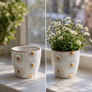 May include: A cylindrical ceramic planter with a glossy white finish and a repeating embossed daisy pattern. Each flower features white petals and a raised yellow centre. The planter has a smooth, rounded rim and a tapered base design.