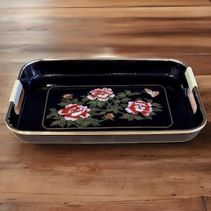 Puede incluir: Bandeja de servir negra con ribete dorado y un diseño floral con peonías rosas y blancas y una mariposa. La bandeja tiene dos asas de color beige claro.
