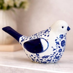 Puede incluir: Figura de pájaro de cerámica en blanco y azul marino. El pájaro tiene un cuerpo blanco con patrones y detalles florales en azul marino. La cola y una zona en forma de ala son de color azul marino liso. El pico también es azul marino.