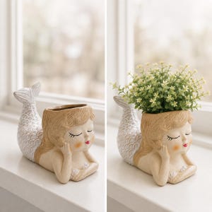 Puede incluir: Jardinera de cerámica beige con forma de sirena, con ojos cerrados, labios rojos y cabello rubio. La sirena está tumbada con la cola levantada. La jardinera está llena de pequeñas flores verdes y blancas. La cola de la sirena es blanca.