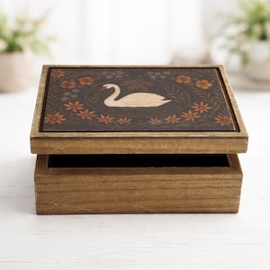 May include: A rectangular wooden box with a dark brown finish. The lid features a central illustration of a white swan surrounded by a circular wreath of orange and brown flowers and green leaves against a dark background.