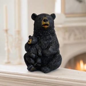 Könnte beinhalten: Eine schwarze Bärenfigur, die einen großen erwachsenen Bären zeigt, der aufrecht sitzt und ein kleines Jungtier im Arm hält. Beide Bären haben strukturiertes Fell, schwarze Körper und beigefarbene Schnauzen mit geöffneten Mäulern.