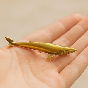 May include: A small, golden whale figurine with detailed fins and a textured body. The whale is approximately 4 inches long, with a pointed snout and a curved tail. The figurine has a vintage, antique appearance.
