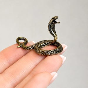 Peut inclure: Une petite figurine en métal de couleur bronze antique représentant un cobra en position enroulée. Le serpent présente un motif texturé en forme d'écailles sur tout le corps, la tête relevée et la gueule ouverte, révélant des crochets détaillés.