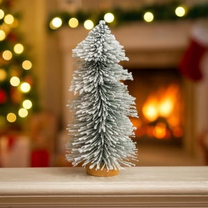May include: A miniature artificial Christmas tree, frosted to resemble snow. The tree is green with white detailing and stands on a circular wooden base. The backdrop includes a fireplace with a warm glow and festive decorations.