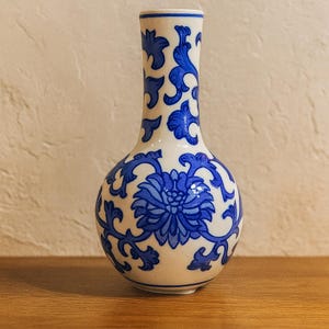 Puede incluir: Un jarrón de cerámica blanco con un diseño floral azul. El jarrón tiene un cuerpo redondeado y un cuello estrecho. El diseño azul presenta una flor grande y patrones arremolinados. El jarrón está sobre una superficie de madera.
