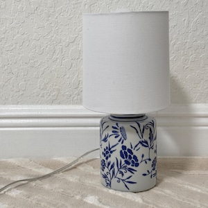 May include: A small table lamp with a white fabric shade. The base is cylindrical, decorated with blue floral patterns on a white background. A power cord extends from the base. The lamp is on a beige rug.