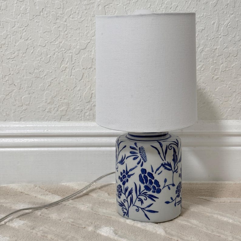 May include: A small table lamp with a white fabric shade. The base is cylindrical, decorated with blue floral patterns on a white background. A power cord extends from the base. The lamp is on a beige rug.