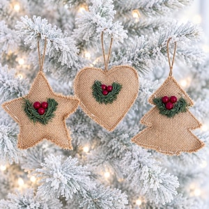 May include: Three hanging ornaments made of tan burlap in the shapes of a star, a heart, and a Christmas tree. Each ornament features a small cluster of green pine needles and three red berries attached to the center with a thin twine loop.