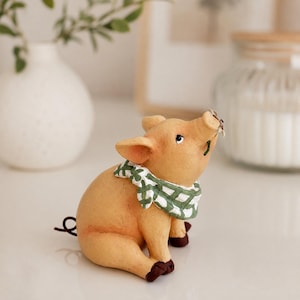 May include: A small, light orange pig figurine in a seated position, wearing a green and white checkered bandana around its neck. The pig has dark brown hooves, a curled tail, and is looking upward with a small flower held in its snout.