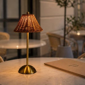 May include: A portable table lamp featuring a conical woven rattan shade and a slender gold-toned metal stem. The lamp has a circular gold base and emits a warm, soft glow from beneath the textured wicker shade, creating a focused light.